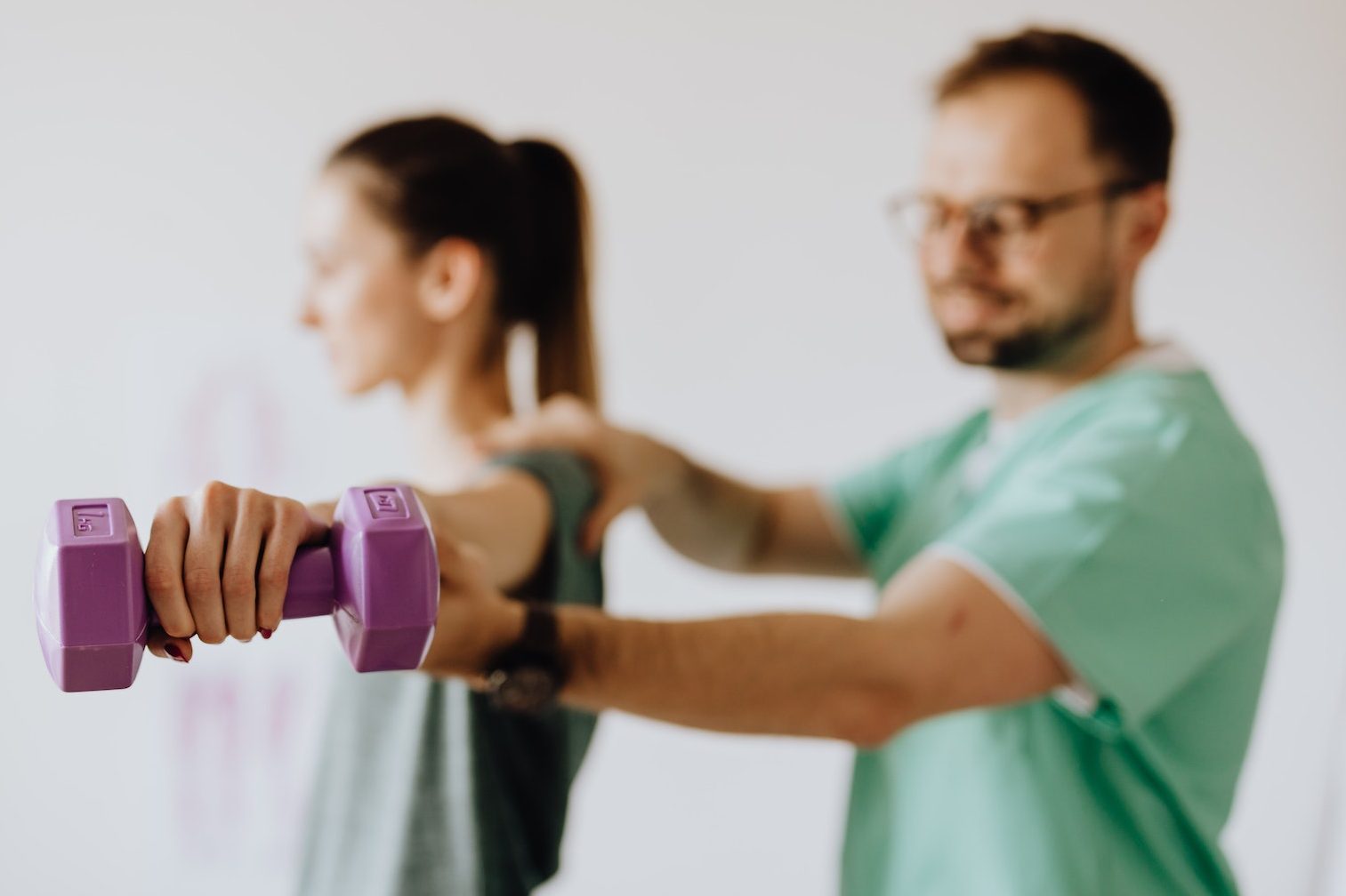 Side view of professional orthopedist in uniform and eyewear helping fit woman reaching arm with dumbbell in doctor office on blurred background