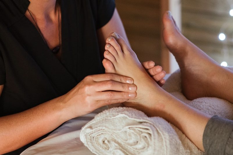 Concentrated female therapist giving foot massage to anonymous patient lying on table with towel during spa procedure on blurred background